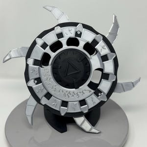 Könnte beinhalten: Ein silbernes und schwarzes 3D-gedrucktes Dekorationsobjekt mit einem dreieckigen Design in der Mitte und einer Reihe von Symbolen am Rand. Das Objekt steht auf einem schwarzen Ständer und einer grauen Oberfläche.