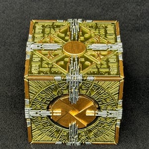 May include: A gold-colored metal cube with intricate designs and a circular opening on each side. The cube has a silver-colored metal frame around each side.