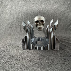 Puede incluir: Una escultura de calavera de metal plateado que levita sobre una base geométrica. La base es de metal y tiene un acabado negro.