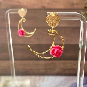 Puede incluir: Pendientes dorados con un diseño de corazón y media luna. Cada luna tiene una pequeña rosa rosa vibrante en el centro. Los pendientes se exhiben en un soporte transparente sobre un fondo borroso.