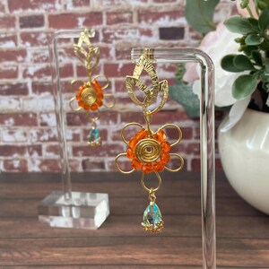Puede incluir: Pendientes de alambre dorado con un diseño de flor naranja y un cristal azul en forma de lágrima. Los pendientes tienen una parte superior en forma de hoja y se exhiben en un soporte transparente. El fondo presenta una pared de ladrillo y flores.