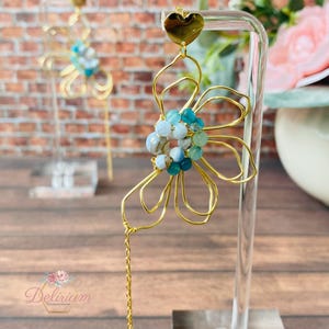 Puede incluir: Pendientes de flor de alambre dorado con cuentas azules y blancas. Los pendientes tienen una parte superior en forma de corazón y una cadena larga que cuelga de la parte inferior.