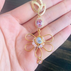 Puede incluir: Un colgante floral dorado con una piedra preciosa ovalada rosa rodeada de pequeñas piedras transparentes. El colgante tiene un diseño de flor con un pequeño dije en forma de corazón rosa en la parte inferior. El colgante se sostiene en una mano.