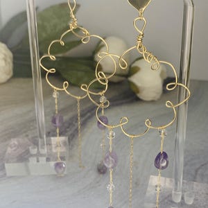 Puede incluir: Pendientes de alambre dorado con un dije de corazón y cuentas colgantes de amatista y cristal transparente.