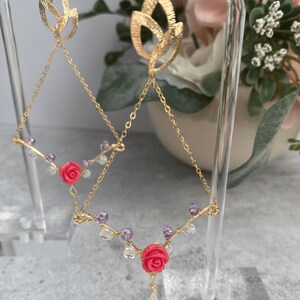 Puede incluir: Un par de pendientes de oro con cadenas delicadas y detalles de flores rosas. Los pendientes presentan un diseño de hoja en la parte superior y cuelgan de una cadena con pequeñas cuentas moradas y transparentes. La parte inferior de los pendientes presenta un dije de flor rosa.