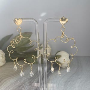 Puede incluir: Pendientes de alambre dorado con parte superior en forma de corazón y un diseño de cristal colgante. Los pendientes tienen forma de nube con un diseño delicado de alambre.
