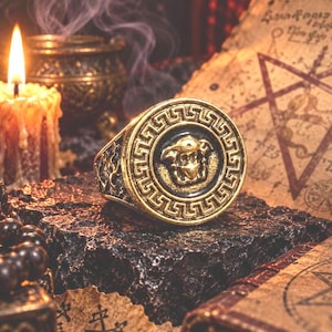 Könnte beinhalten: Ein goldfarbener Metallring mit einer kreisförmigen Vorderseite, die ein erhabenes Medusenhaupt-Emblem zeigt, umgeben von einem Mäander-Muster. Der Ringkörper weist an den Seiten komplizierte Reliefschnitzereien auf einem dunklen Untergrund auf.