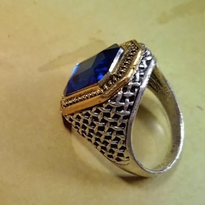 May include: A silver ring with a blue gemstone set in a gold frame. The ring has a woven band.
