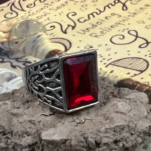 Puede incluir: Un anillo plateado con una gran gema rectangular de color rojo intenso. El anillo tiene un intrincado diseño calado en la banda. El anillo se exhibe sobre un trozo de madera.