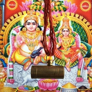 May include: A gold-coloured metal cylinder pendant hanging from a red string. The pendant is in front of a colourful painting of two deities, a man and a woman, sitting on a throne. The painting has a floral design and the word "சக்தி" written in Tamil.