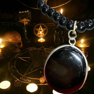 May include: A black onyx pendant necklace with a silver chain and black beads. The necklace is hanging over a pentagram and candles on a black altar.