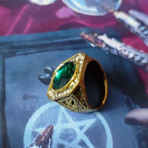 May include: A gold ring with a large, oval-shaped green gemstone surrounded by smaller clear gemstones. The ring has a detailed design on the band.