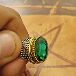 Könnte beinhalten: Silberfarbener Ring mit goldfarbenem Rahmen und einem großen, oval geschliffenen, smaragdgrünen Edelstein. Das Ringband hat ein strukturiertes, gepunktetes Muster. Der Edelstein reflektiert das Licht.