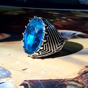 May include: A silver-toned ring featuring a large, oval-cut, bright blue gemstone. The ring has a detailed, geometric design on the band and around the gem, creating a striking contrast. The gemstone is the focal point, catching the light and adding a touch of elegance.