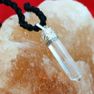 May include: A clear quartz crystal pendant with a silver setting, hanging from a black cord. The pendant is resting on a light brown surface.