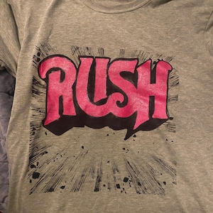 May include: A grey t-shirt with the word "RUSH" in a pink, graffiti-style font with a black outline. The design includes a black burst and splatter effect. The shirt is made of a soft material.