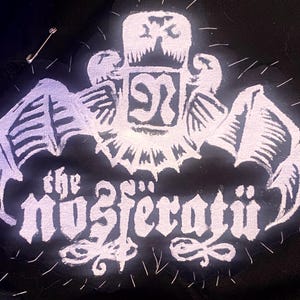 May include: White embroidered patch with a gothic font design that reads "the nosferatu". The design includes a stylized bat wing and a crown with a letter "N" inside.