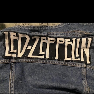 Puede incluir: Chaqueta vaquera azul con un parche rectangular grande cosido en la espalda. El parche es negro con el texto blanco "LED ZEPPELIN" en una fuente audaz y estilizada.