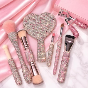 Puede incluir: Una colección de herramientas de maquillaje glamurosas, que incluyen brochas, un espejo en forma de corazón y un rizador de pestañas. Los mangos y el espejo están incrustados con brillantes pedrería en tonos rosa y transparente. Las brochas tienen cerdas suaves de color rosa.
