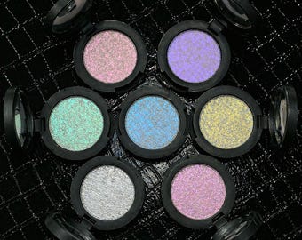 May include: Seven open, black compacts of iridescent eyeshadow in various colors, including pink, purple, green, blue, yellow, and silver. The eyeshadows have a shimmering, glittery finish. The compacts are arranged on a black, textured surface.