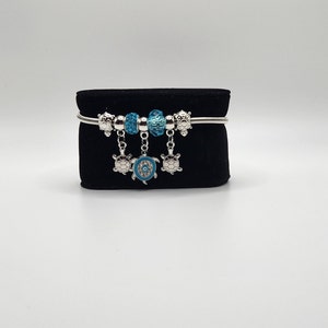 Op de afbeelding: Een zilveren armband met blauwe en zilveren schildpadbedels. De armband heeft een zilveren ketting en is versierd met blauwe en zilveren kralen.