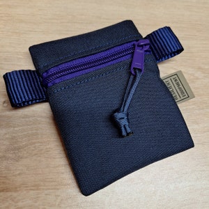 Op de afbeelding: Een klein, donkerblauw ritsvakje met een paarse rits en een donkerblauwe band. Het vakje heeft een klein label met de tekst "RESILIENT EQUIPMENT".