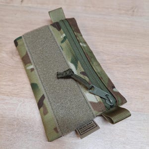 Könnte beinhalten: Eine kleine, grün-braune Camouflage-Tasche mit Reißverschluss, Klettverschluss und Zuglasche. Die Tasche hat ein Etikett mit der Aufschrift "Resilient Equipment".