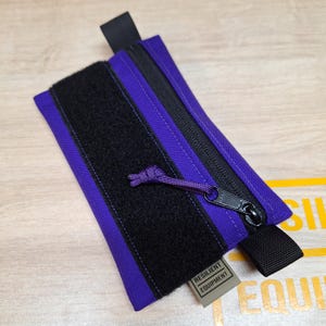 May include: A purple and black zipped pouch with a black Velcro strip and a black pull tab. The pouch has a small tag that reads "Resilient Equipment". The zipper pull is purple.