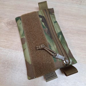 Könnte beinhalten: Eine kleine, camouflagefarbene Reißverschlusstasche mit einem braunen Klettverschluss und einem braunen Zugband. Die Tasche hat ein Etikett mit der Aufschrift "RESILIENT EQUIPMENT".