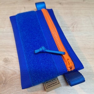 Op de afbeelding: Een kleine blauwe ritszak met een oranje rits en een blauwe band. De zak heeft een klittenbandsluiting en een label met de tekst "RESILIENT EQUIPMENT".