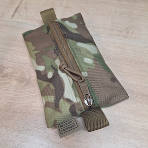 Könnte beinhalten: Eine kleine, camouflagefarbene Reißverschlusstasche mit einem braunen Riemen und einem Etikett mit der Aufschrift "RESILIENT EQUIPMENT".