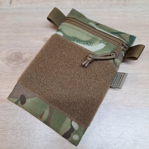 Könnte beinhalten: Eine kleine, camouflagefarbene Tasche mit Reißverschluss und Klettverschluss. Die Tasche hat einen braunen Riemen und einen braunen Zugverschluss. Die Tasche ist mit "Resilient Equipment" beschriftet.