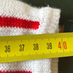 May include: Close-up of a white knitted fabric with two red stripes. A yellow measuring tape shows a length of approximately 15 inches. The fabric appears to be part of a garment.