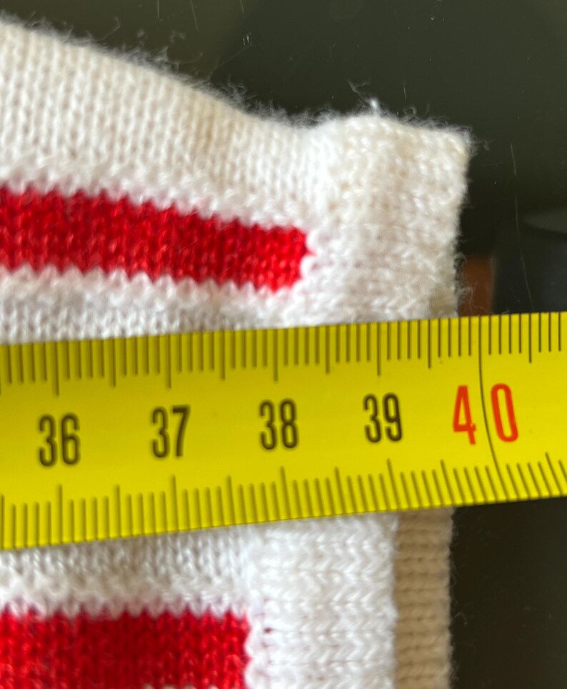 May include: Close-up of a white knitted fabric with two red stripes. A yellow measuring tape shows a length of approximately 15 inches. The fabric appears to be part of a garment.