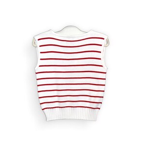 May include: White sleeveless sweater with horizontal red stripes. The sweater is hanging on a white hanger. The bottom hem and neckline are solid white.