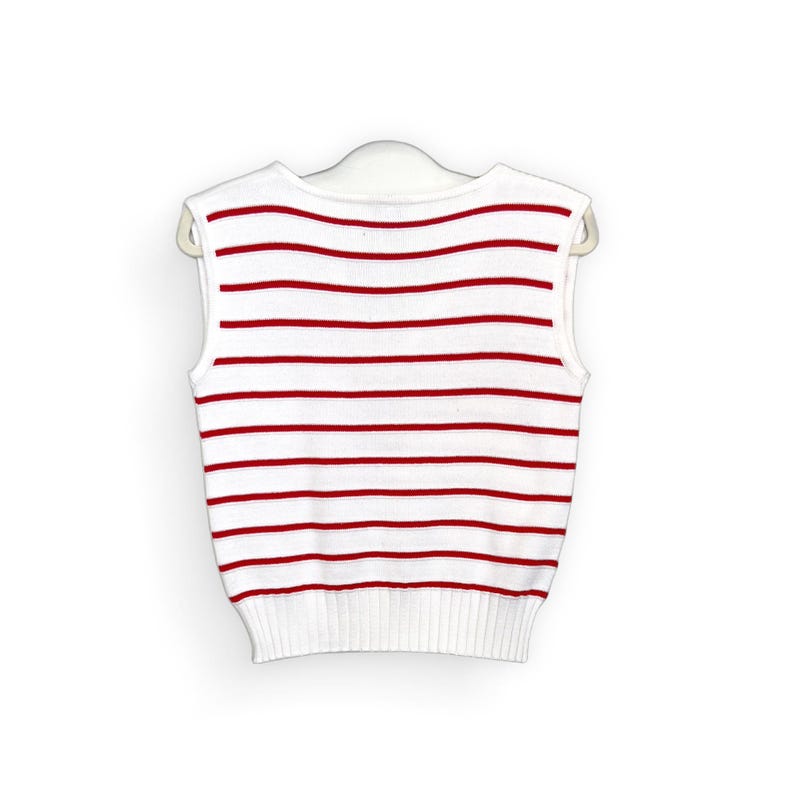 May include: White sleeveless sweater with horizontal red stripes. The sweater is hanging on a white hanger. The bottom hem and neckline are solid white.