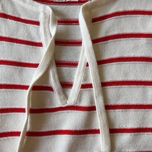 May include: A close-up of a white and red striped sweater. The sweater has a V-neck with a drawstring and the brand name "courreges paris" is visible on a tag.