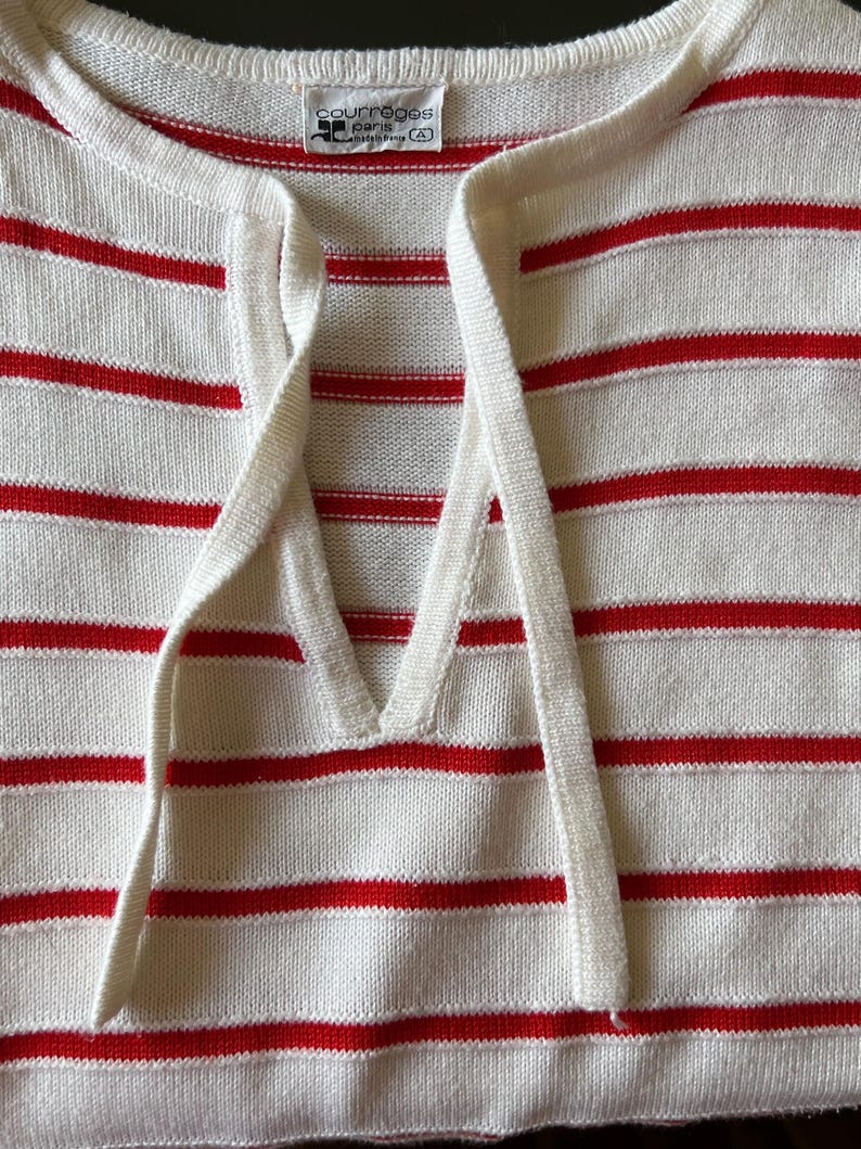 May include: A close-up of a white and red striped sweater. The sweater has a V-neck with a drawstring and the brand name "courreges paris" is visible on a tag.
