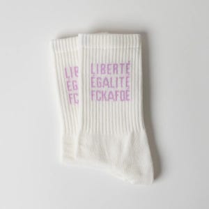 Könnte beinhalten: Ein Paar weiße Socken mit Rippenstruktur und dem Text "LIBERTÉ ÉGALITÉ FCKAFDE", der in helllila Farbe auf der Seite aufgedruckt ist. Die Socken haben eine Standardlänge und ein schlichtes Design mit einer klaren, gut lesbaren Schriftart.
