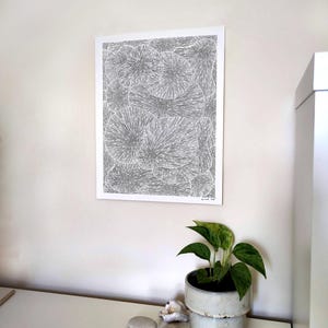May include: A framed black and white art print with a detailed, abstract design of circular shapes. The print hangs on a white wall above a white table. A small potted plant and decorative stones are on the table.