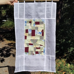 May include: A rectangular wall hanging featuring a central patchwork panel of assorted fabric scraps in shades of burgundy, sage green, yellow, and blue floral patterns, framed by a sheer white border with vertical and horizontal white fabric strips.
