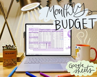 May include: A laptop computer displaying a Google Sheets spreadsheet with a monthly budget template. The spreadsheet is titled "Monthly Budget" and has a purple color scheme. The laptop is on a wooden desk with a mug, a plant, and pens.