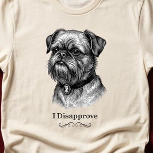 May include: A cream-colored short-sleeve t-shirt featuring a black and white ink-style illustration of a Brussels Griffon dog wearing a collar with a cameo pendant. Below the dog, the text reads I Disapprove in a serif font with a flourish.