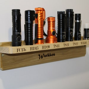 Puede incluir: Una estantería de pared de madera con una variedad de linternas, incluyendo modelos negros, naranja y plateados. La estantería está etiquetada con los nombres de las linternas, incluyendo FC13s, HD15, HD20, TS11, TS21, TS22, WK03 y WK04. La estantería también está etiquetada con la palabra "Wurkkos".