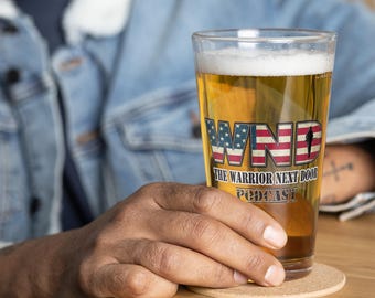 May include: A glass of beer with a white foam head. The glass has a graphic of an American flag with the text "WND The Warrior Next Door Podcast".