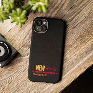 May include: A black smartphone case with a speckled texture and a graphic design featuring the yellow text NEW, three red skull icons, the red text ACHIEVEMENT, and the yellow text CRAWLER # 4122 printed on the lower portion of the back surface.