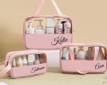 Peut inclure: Trois trousses de maquillage roses avec des côtés transparents et des poignées. Elles contiennent divers produits de beauté. La plus grande porte le nom "Katie", les plus petites "Tatiana" et "Ana".