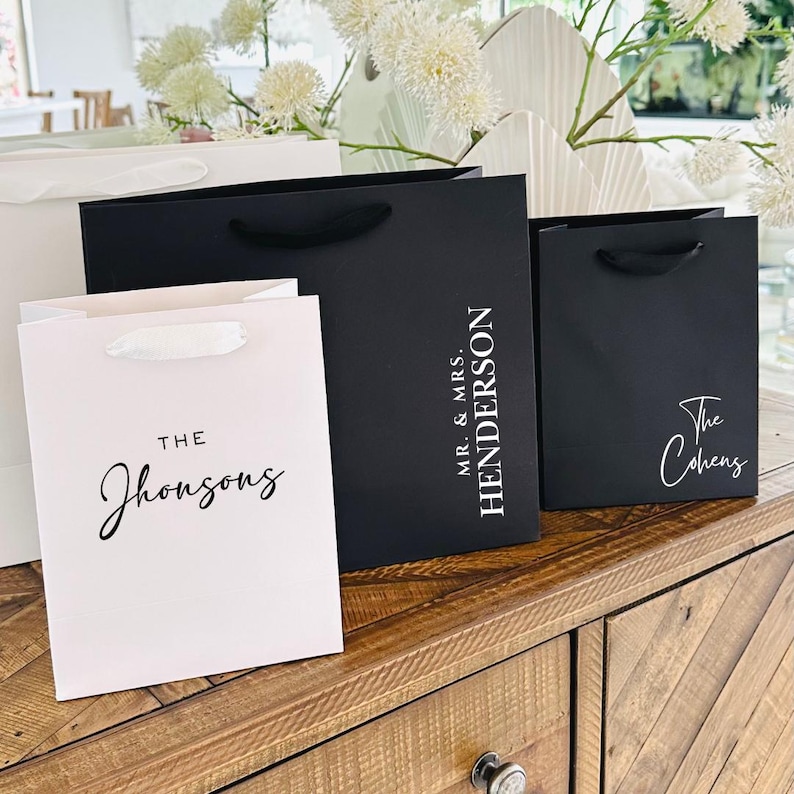 Pu&ograve; includere: Sacchetti regalo bianchi e neri con testo personalizzato. Le borse hanno manici coordinati e sono esposte su una superficie di legno. Il testo include "The Jhonsons", "Mr. & Mrs. Henderson" e "The Cohens".