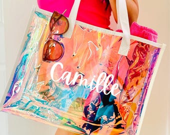 May include: A clear, iridescent tote bag with white handles and trim. The bag features the name "Camille" in white script. Brown sunglasses are clipped to the top, and a pink towel is visible inside. The bag is held by a person wearing a pink top.