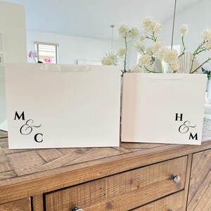 Pu&ograve; includere: Due sacchetti regalo bianchi con manici in raso sono su una superficie di legno. Le borse hanno iniziali nere e una e commerciale. Una borsa mostra "M & C", l'altra "H & M".